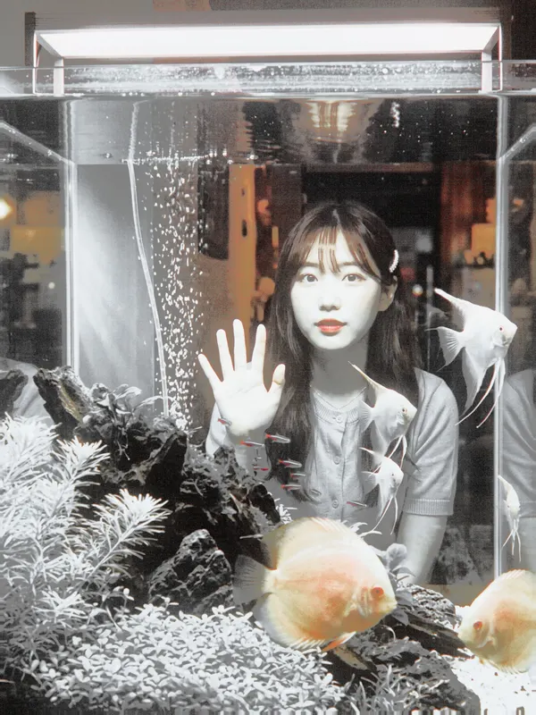 A girl behind a fish tank in black and white, with only the fish left orange-red.
